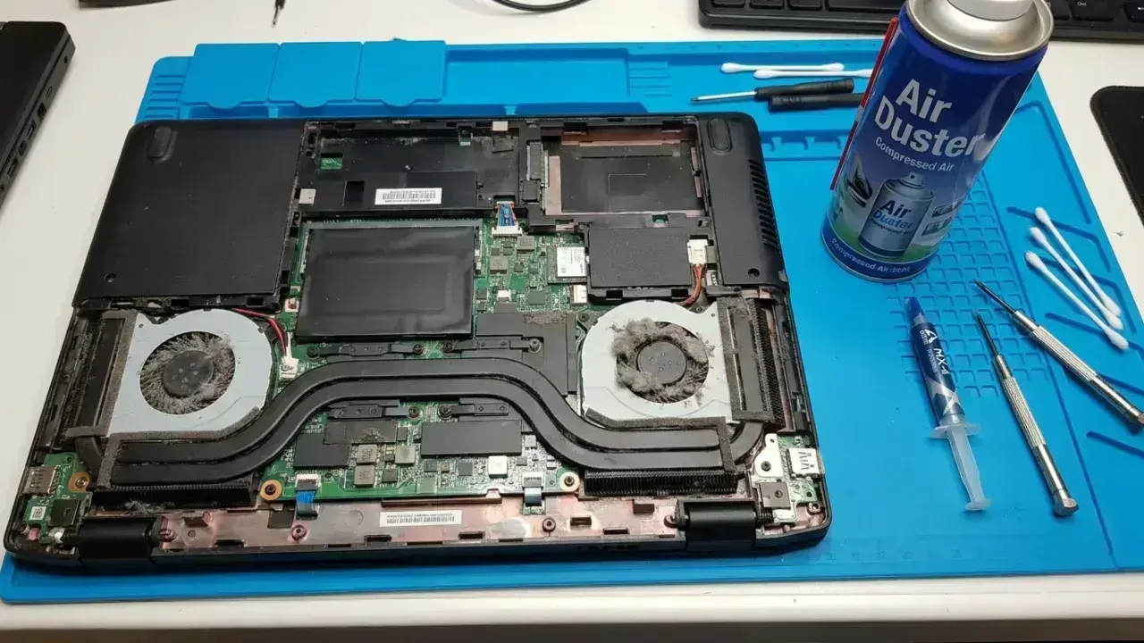 Disassembled laptop with dusty cooling fans next to compressed air duster, thermal paste, and screwdrivers for maintenance.